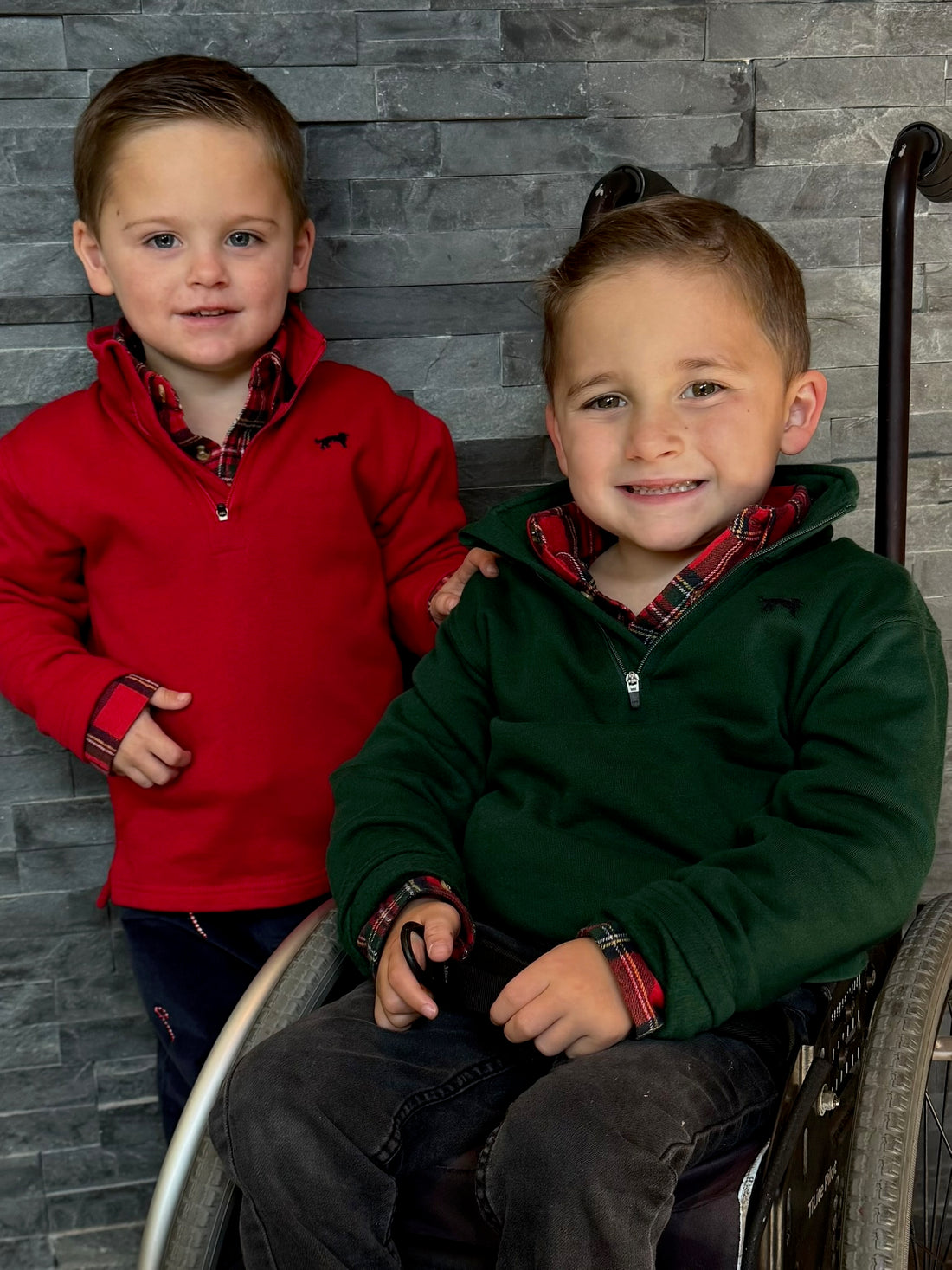 Two young boys, one in a wheelchair and one standing, against a gray brick wall.