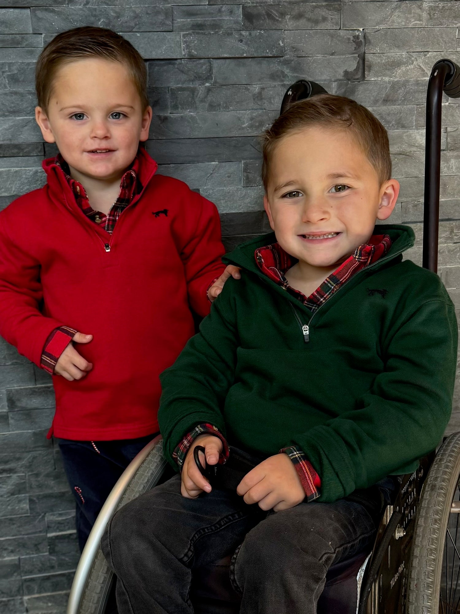 Two young boys, one in a wheelchair and one standing, against a gray brick wall.