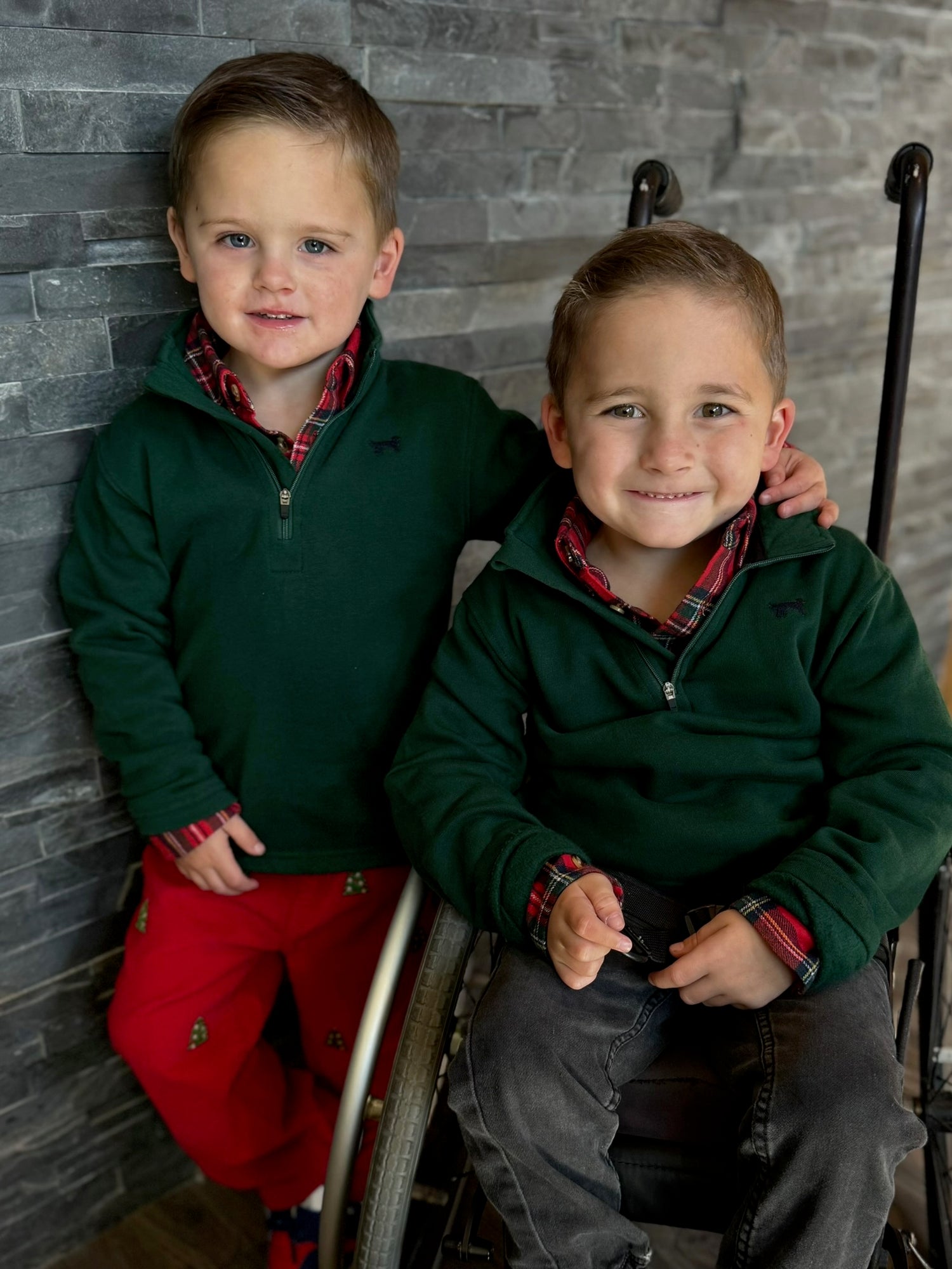 Two young boys wearing matching green jackets and red plaid shirts against a stone wall.