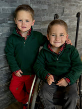 Two young boys wearing matching green jackets and red plaid shirts against a stone wall.