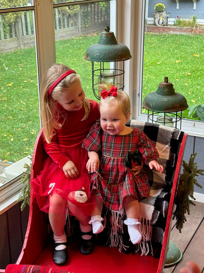 Two young girls in red dresses sitting on a red bench with a window in the background.