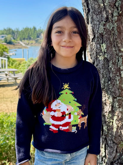 Child wearing a Christmas-themed sweater with Santa Claus and a tree, standing next to a tree outdoors.
