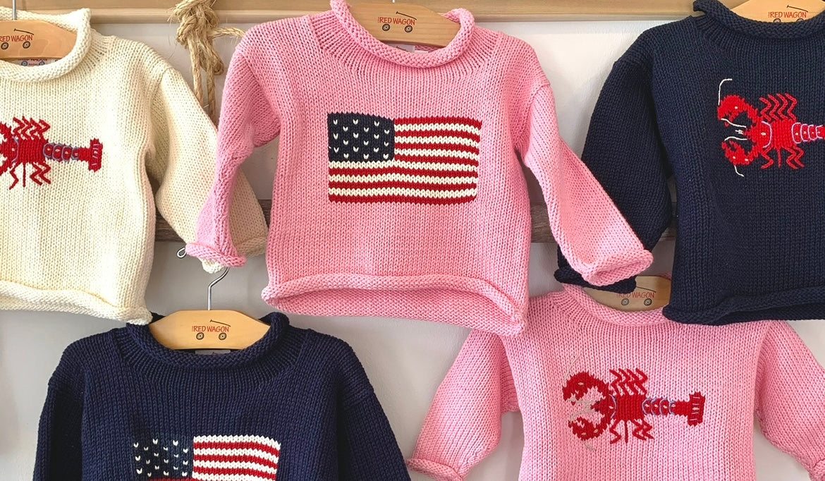 image shows pink flag sweater, navy lobster sweater, ivory lobster sweater, navy flag sweater and pink lobster sweater all hanging on the wall