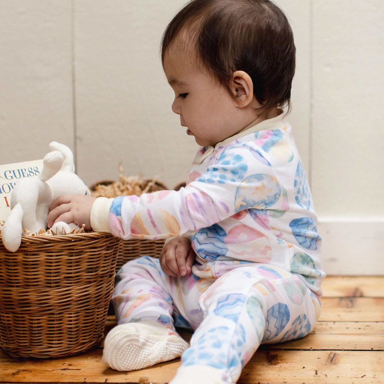 Baby in a colorful outfit sitting next to a wicker basket with toys on a wooden floor.