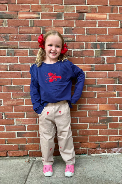Young girl wearing a blue sweatshirt with a red design, beige pants, and pink shoes against a brick wall.