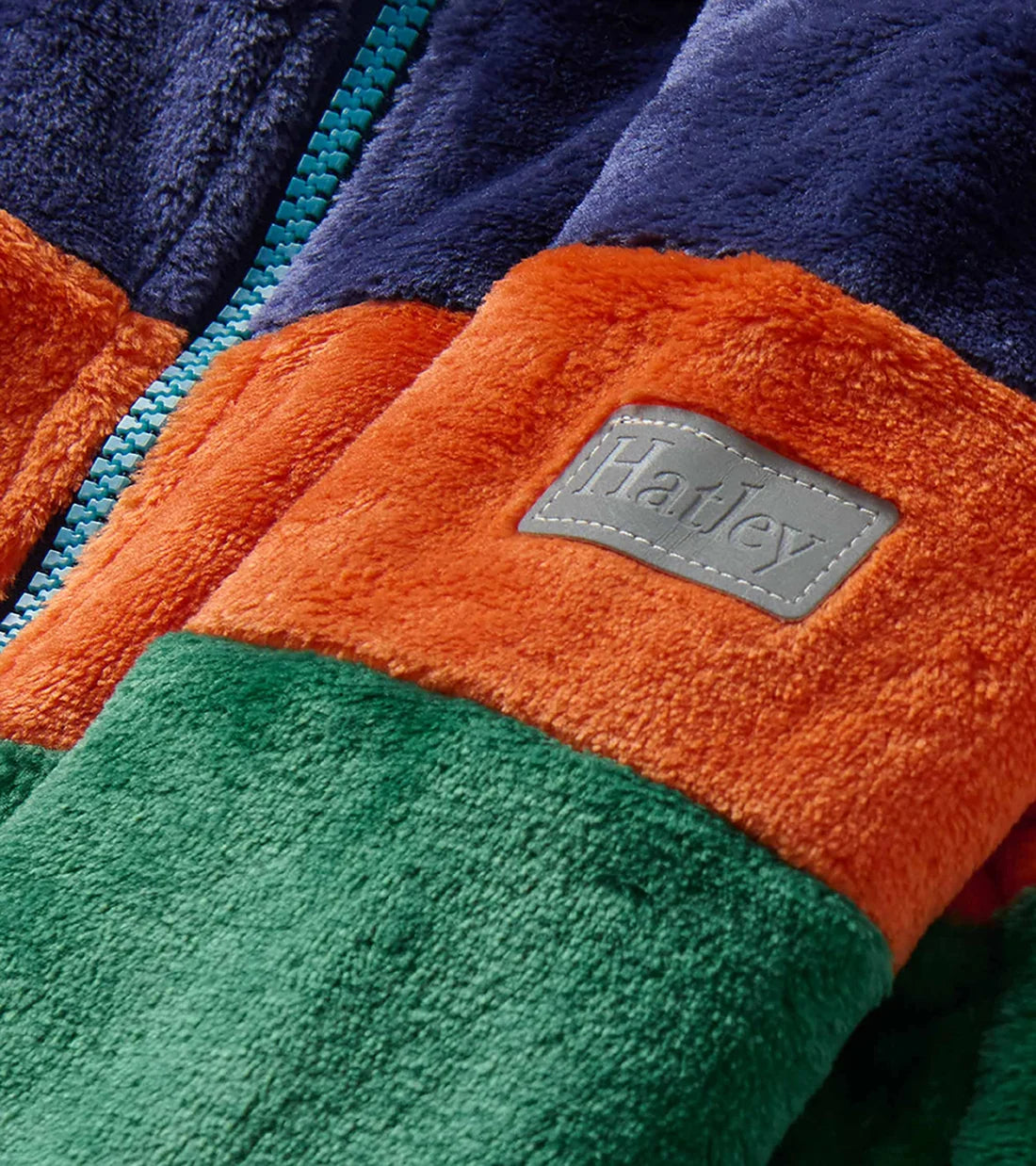 Close-up of a plush fleece zip jacket with color-blocked sections in navy, orange, and green, featuring a gray patch with the embossed brand name Hatley.