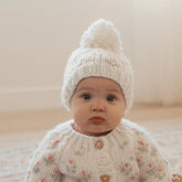 Baby wearing a white knitted outfit with a matching hat on a neutral background
