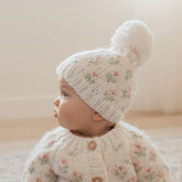 Baby wearing a floral knitted outfit with a white hat featuring a pom-pom on a neutral background