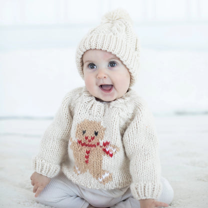 Baby wearing a cream knitted sweater with a gingerbread design and matching hat on a white background