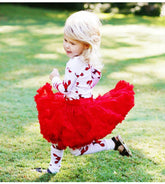 A toddler girl with blonde hair is running on a green grass lawn, wearing a white long-sleeve top and leggings patterned with red leaves, paired with a voluminous bright red tulle tutu skirt.