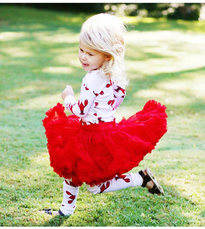 A toddler girl with blonde hair is running on a green grass lawn, wearing a white long-sleeve top and leggings patterned with red leaves, paired with a voluminous bright red tulle tutu skirt.
