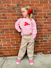 Young girl in pink sweatshirt with red lobster design and beige pants standing against a brick wall.
