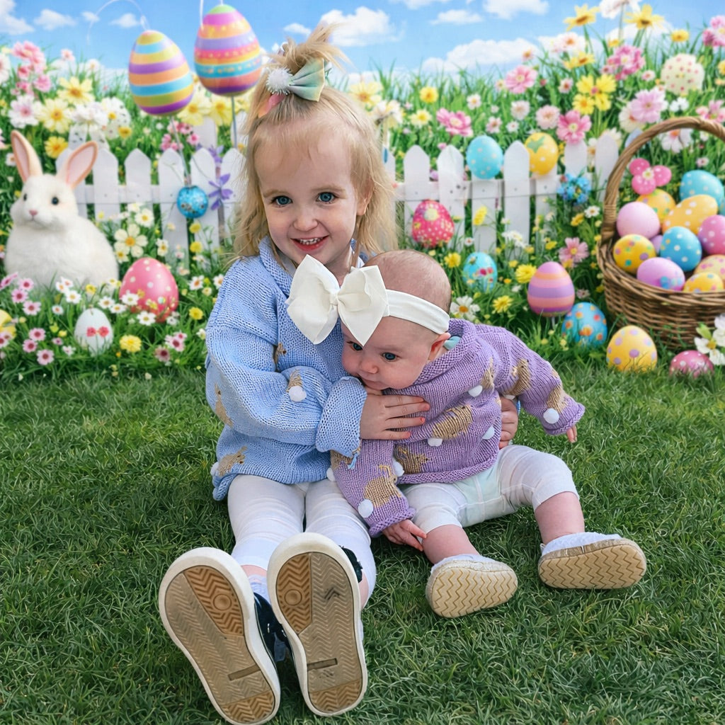 Two young children sitting on grass in front of a white picket fence decorated with Easter eggs, flowers, a bunny, and a basket. The older child wears a light blue knit sweater, white leggings, and black-and-white shoes, while the younger child wears a lavender knit sweater, white leggings, and tan shoes.