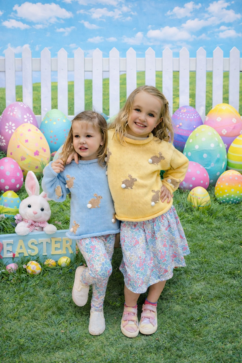 Two children in Easter-themed outfits standing in front of colorful Easter eggs and a white picket fence.