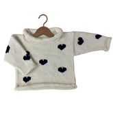 A cream-colored hand-knit baby sweater with black heart patterns, displayed on a wooden hanger against a plain white background.