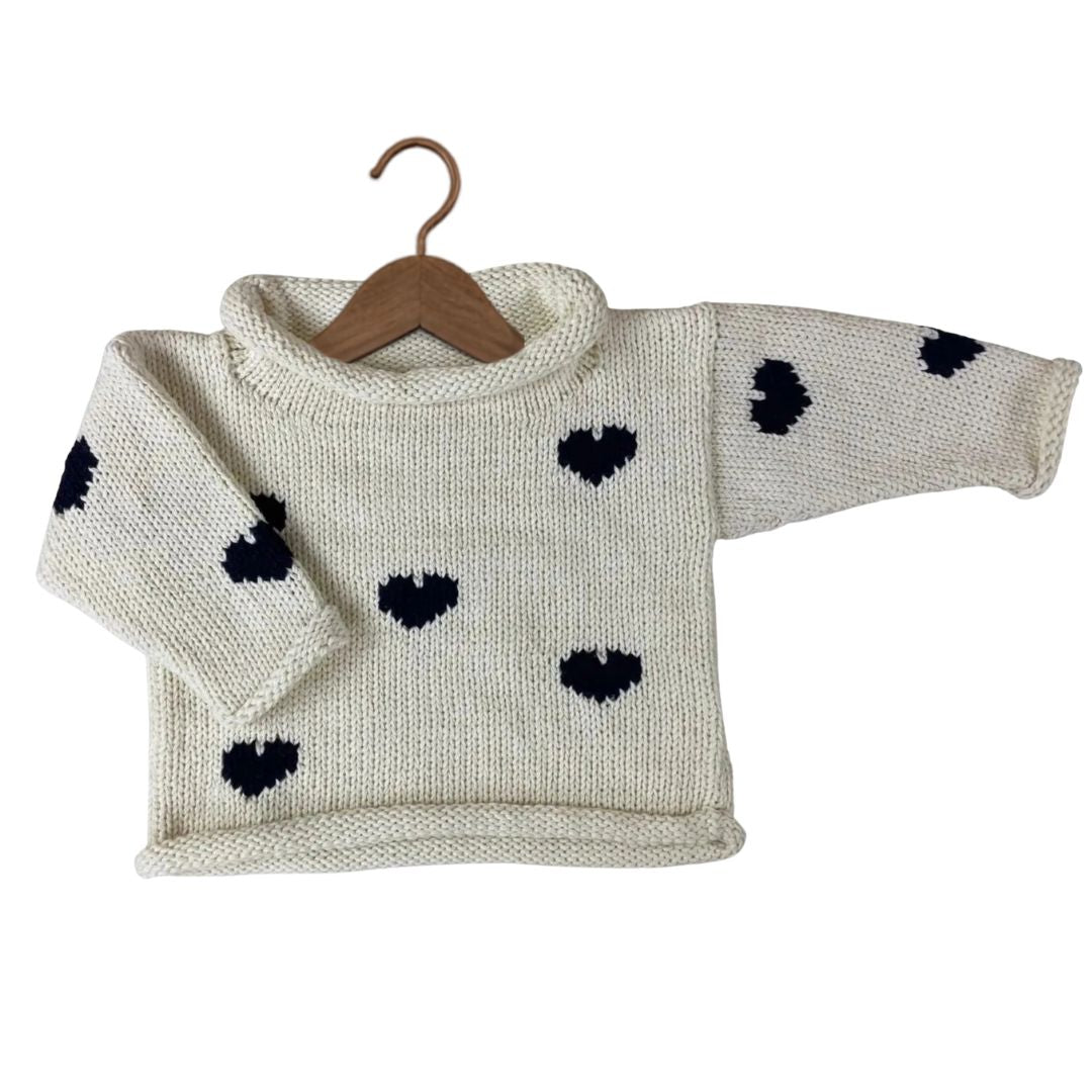 A cream-colored hand-knit baby sweater with black heart patterns, displayed on a wooden hanger against a plain white background.