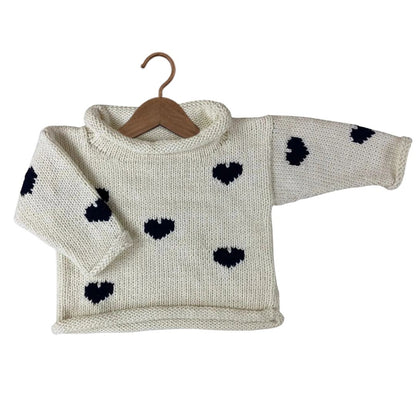 A cream-colored hand-knit baby sweater with black heart patterns, displayed on a wooden hanger against a plain white background.
