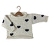 A cream-colored hand-knit baby sweater with black heart patterns, displayed on a wooden hanger against a plain white background.