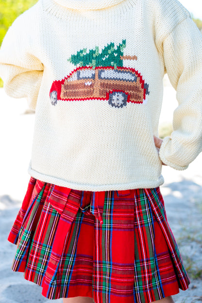 Child wearing a sweater with a Christmas tree and car design and a red plaid skirt.