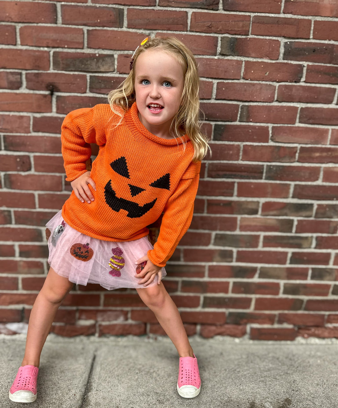 Child wearing an orange sweater with a pumpkin face design, standing against a brick wall.