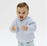 A baby wearing a light blue fuzzy zip-up hoodie and light blue patterned pants, standing against a plain white studio background.