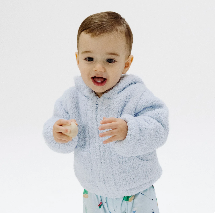 A baby wearing a light blue fuzzy zip-up hoodie and light blue patterned pants, standing against a plain white studio background.