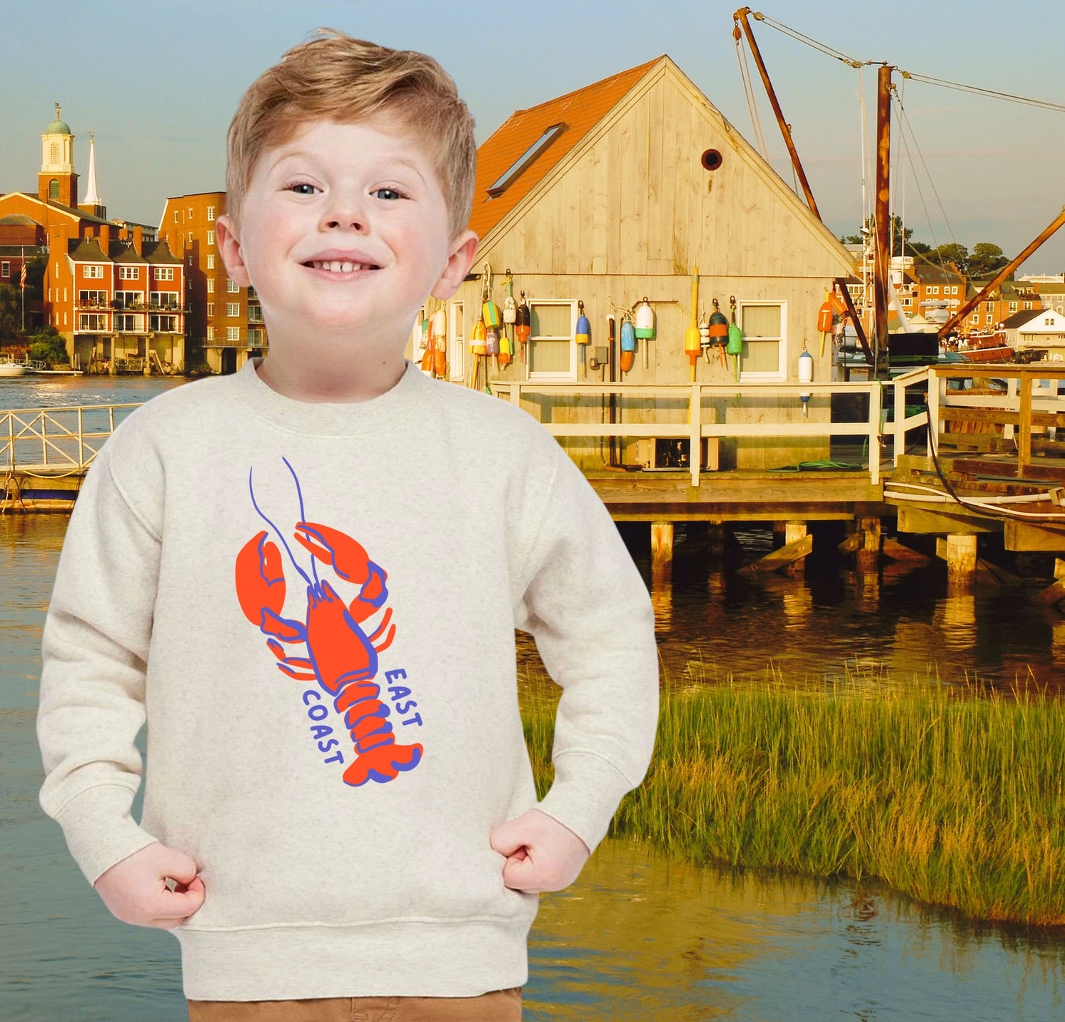 Child wearing a sweater with a lobster design in front of a waterfront scene with buildings and a dock.