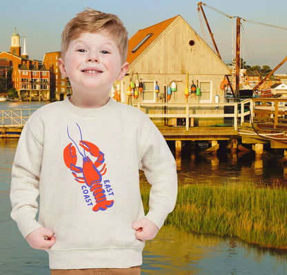 Child wearing a sweater with a lobster design in front of a waterfront scene with buildings and a dock.