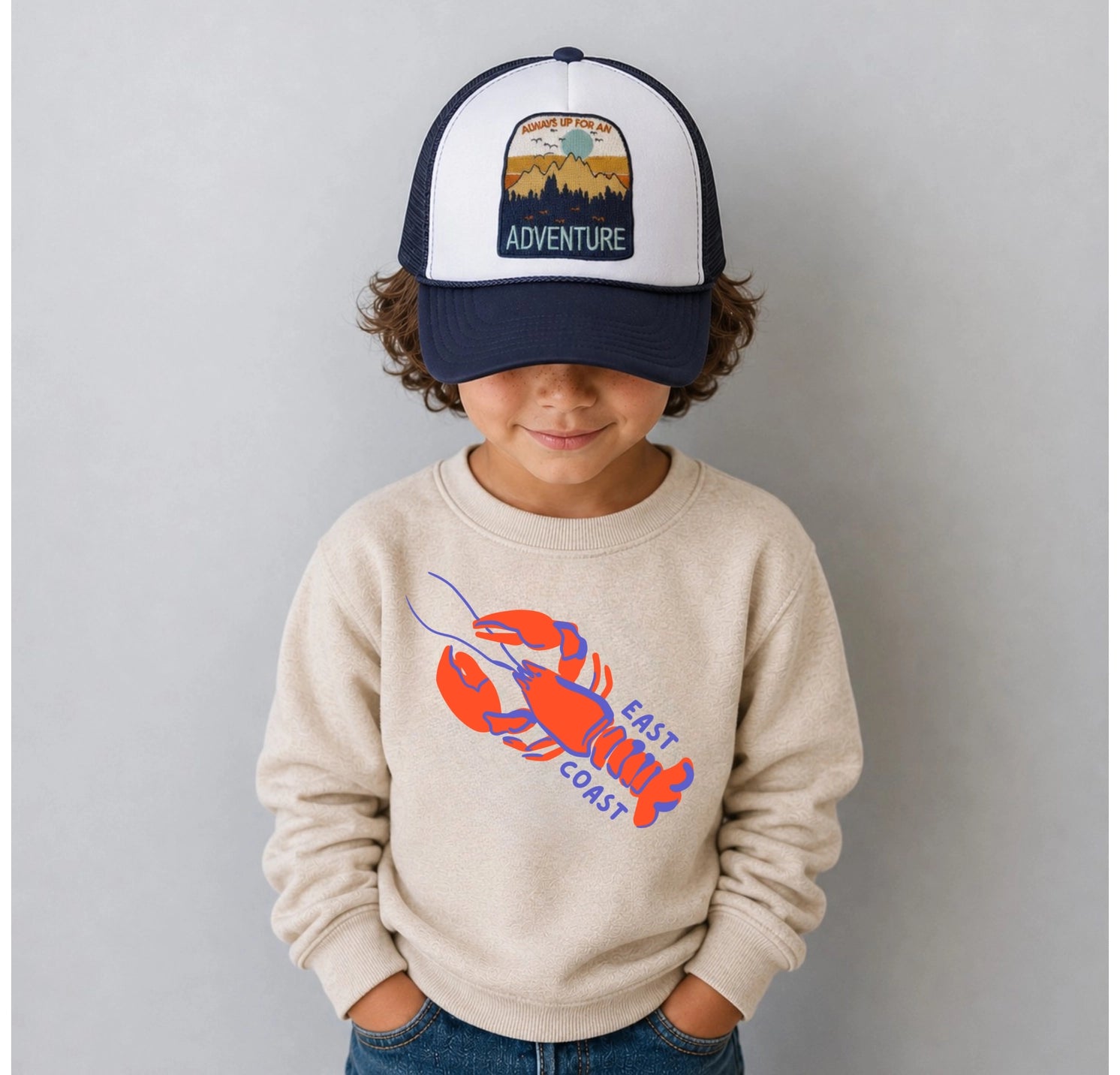 Child wearing a cap with a mountain design and a sweatshirt with a lobster graphic on a plain background