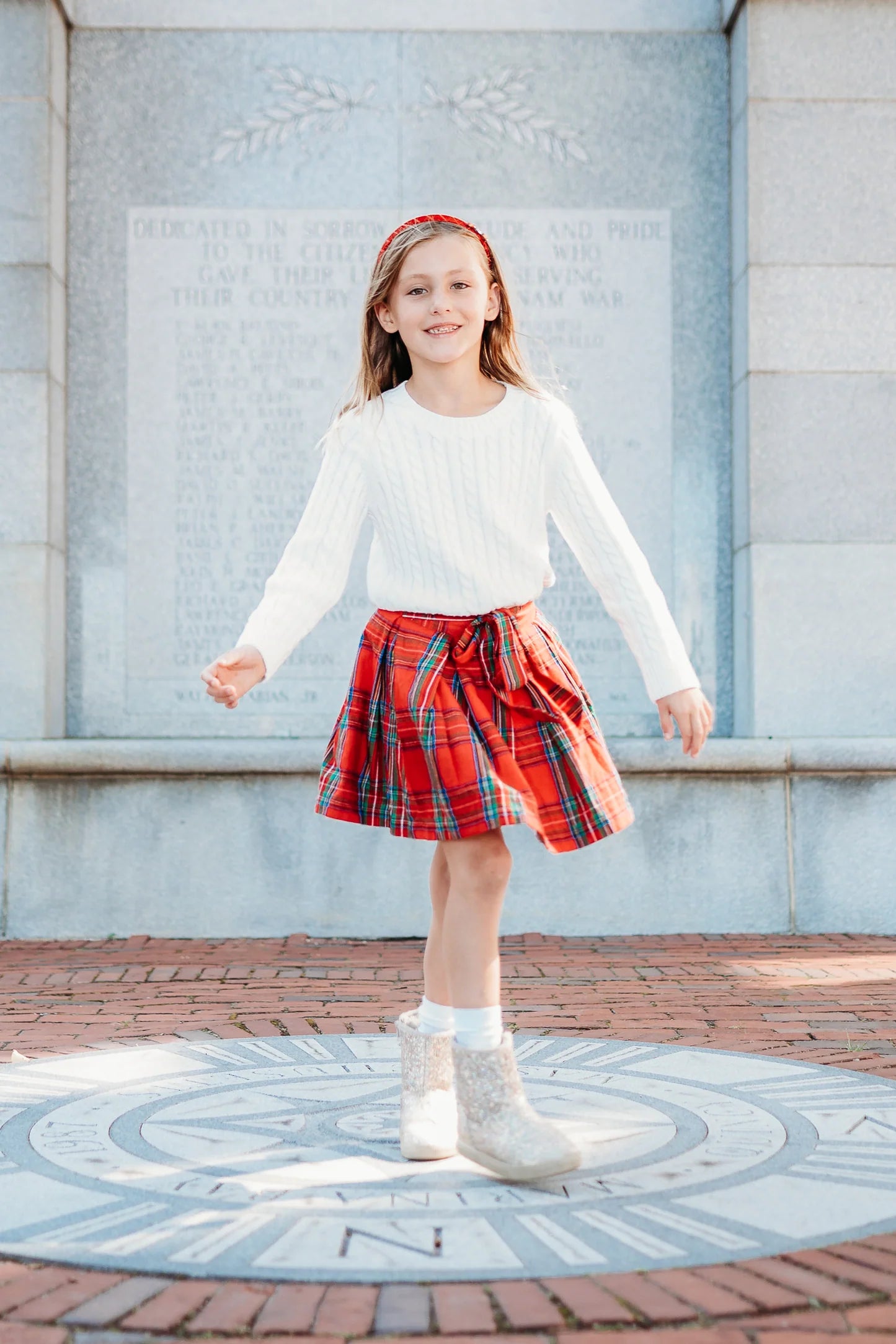 Young girl in a white sweater and red plaid skirt standing on a decorative pavement.