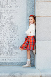 Young girl in a red plaid skirt and white sweater standing against a wall with engraved names.