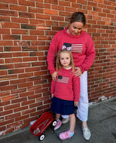 Woman and young girl wearing matching red sweatshirts with American flag design against a brick wall.