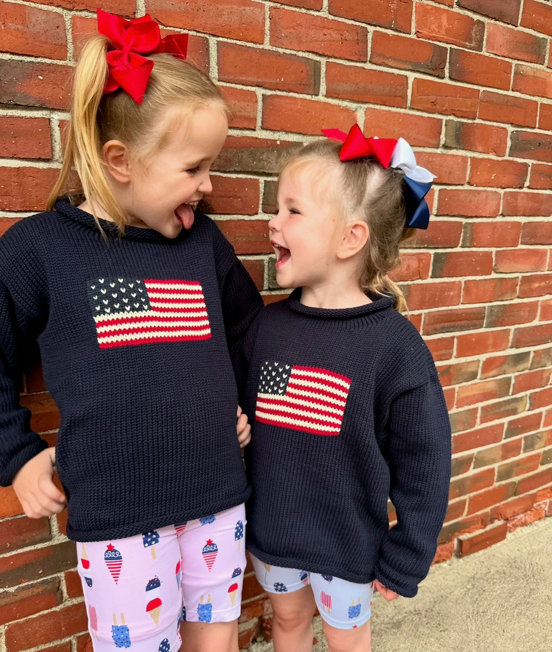 two girls wearing flag sweaters with bows in their hair smiling at each other