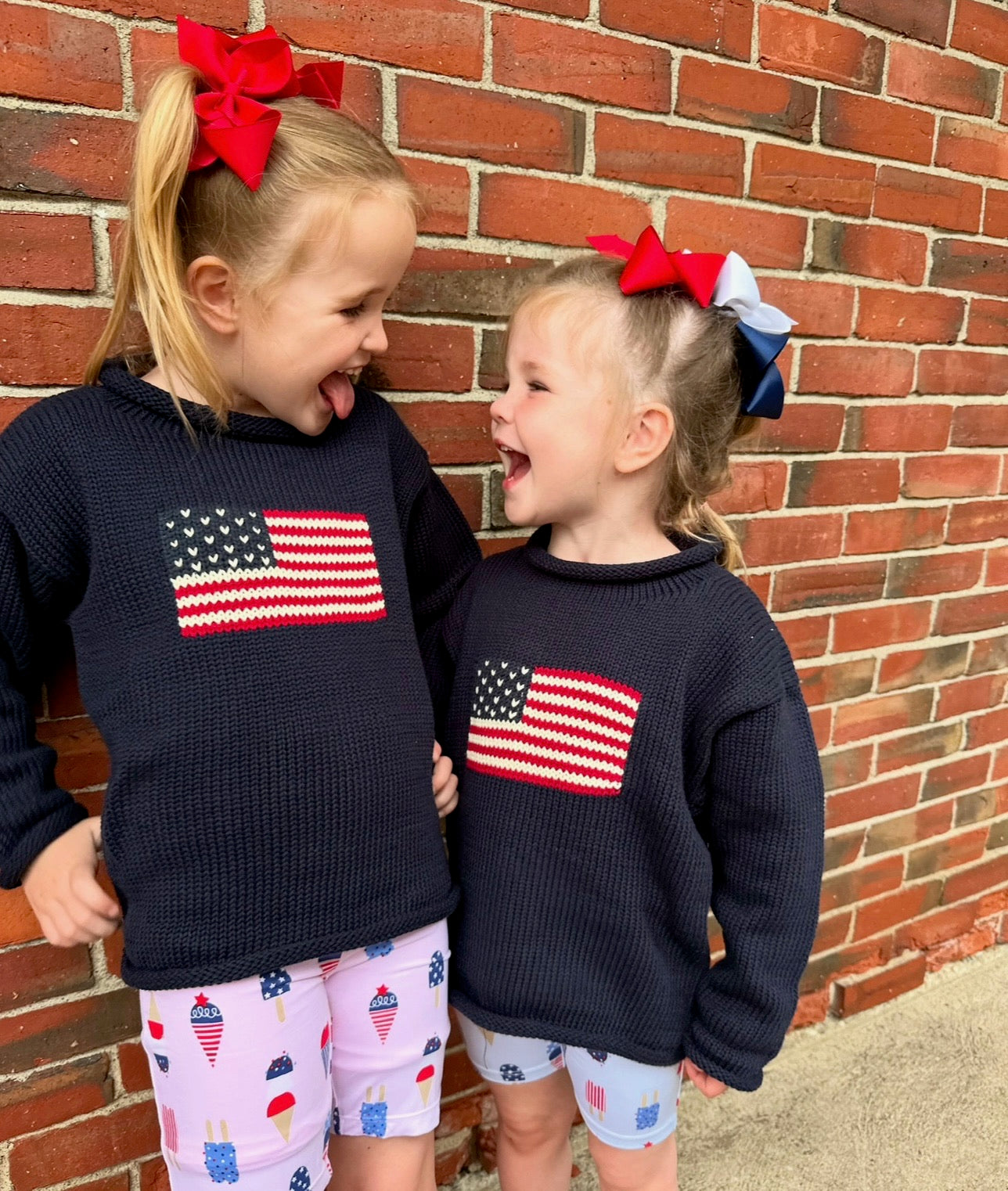 two girls wearing flag sweaters with bows in their hair smiling at each other