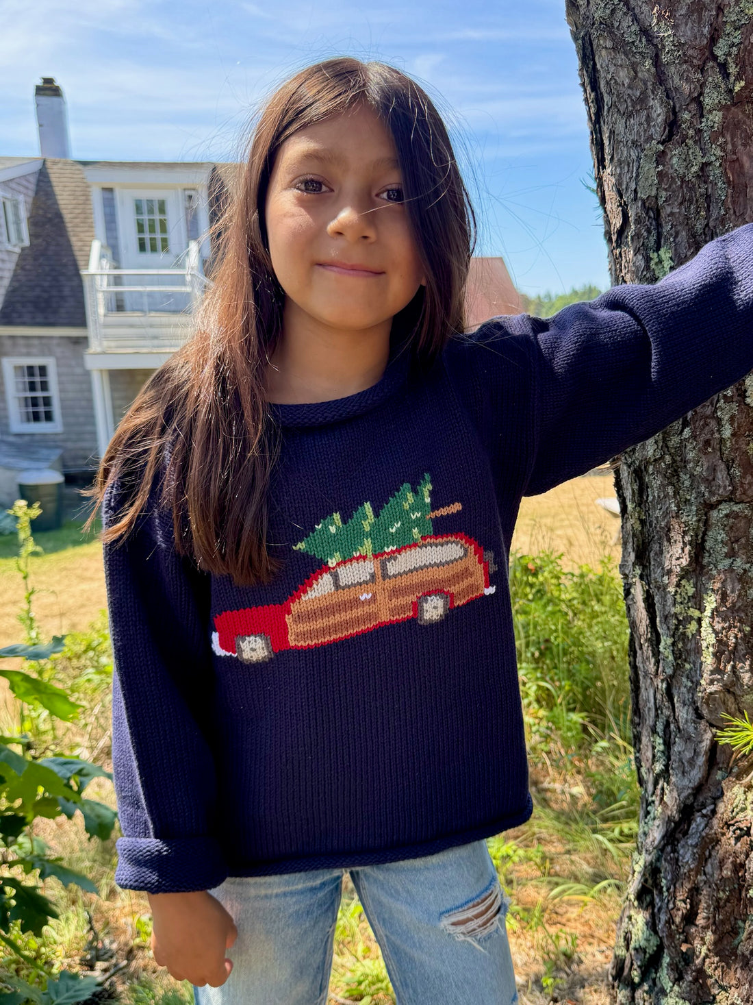 Child wearing a navy sweater with a red car design, standing outdoors near a tree.