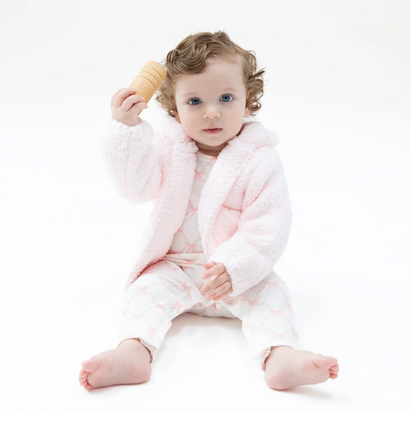 A baby sitting on a white studio background wearing a fluffy pale pink and white patterned romper, holding a small wooden toy near their head.
