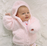A baby wearing a fluffy light pink hooded jacket, lying on a white faux fur blanket.