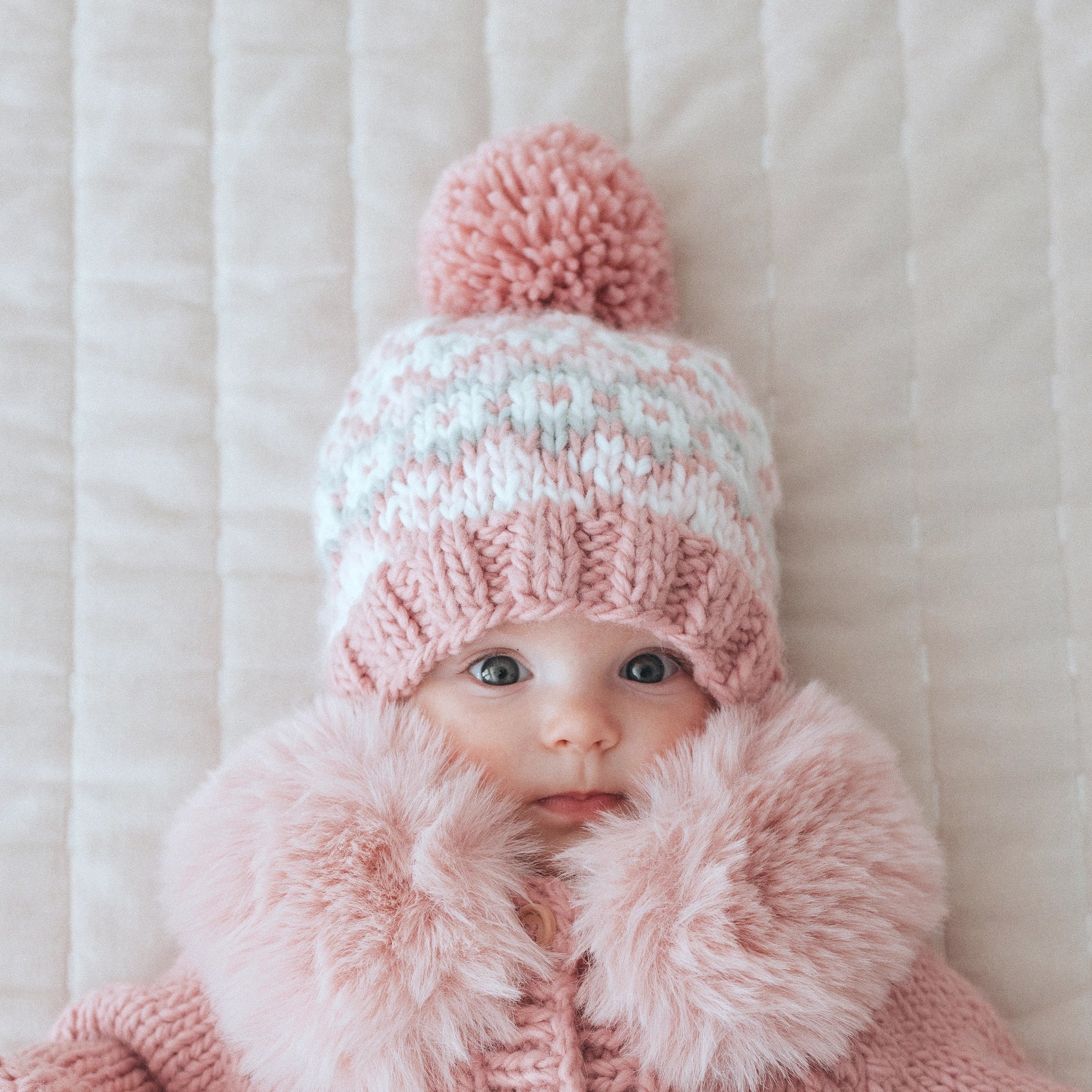 Baby wearing a pink knitted hat with a pom-pom and matching pink coat on a white background