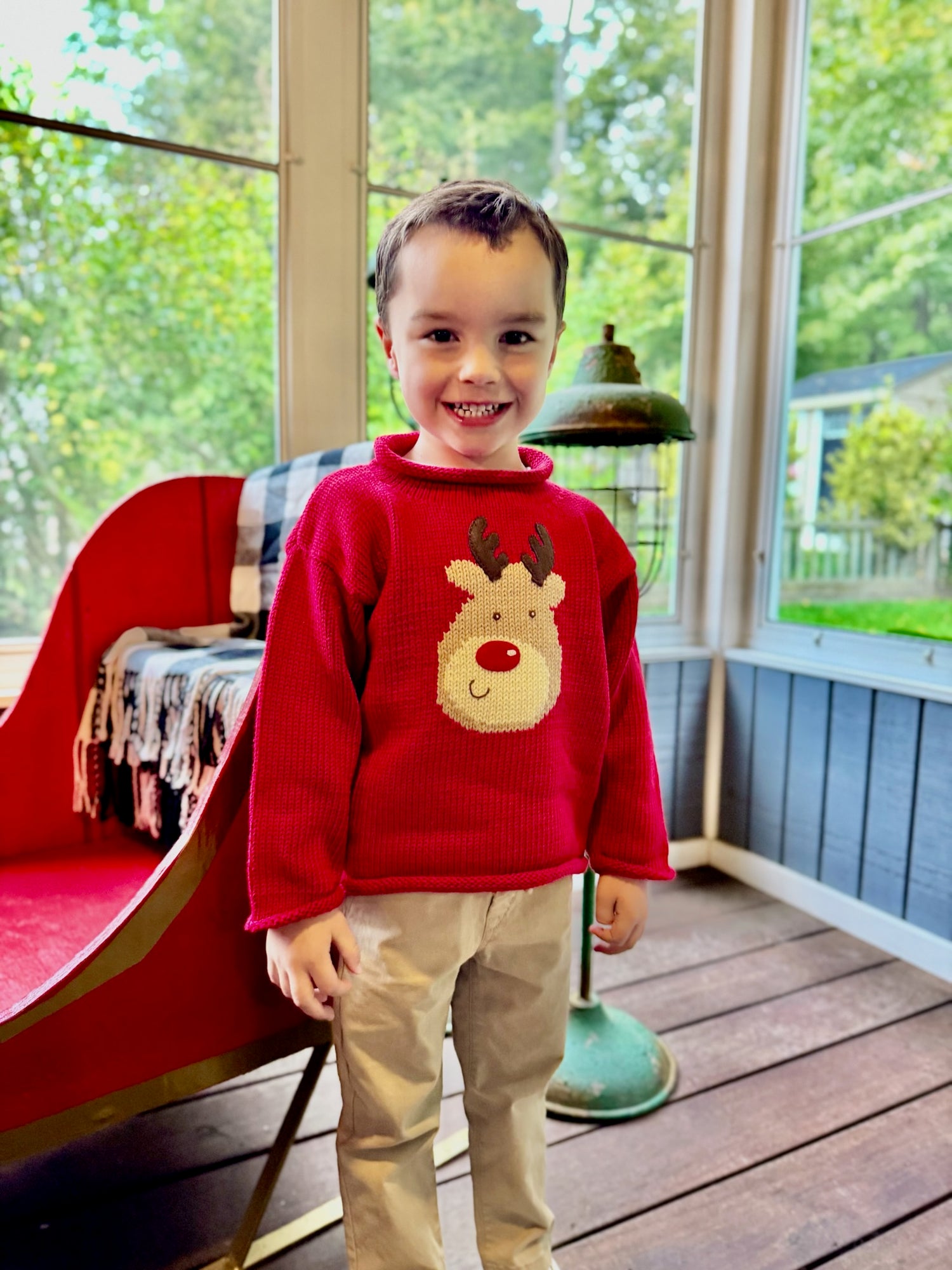 Child wearing a red sweater with a reindeer design on a wooden deck.