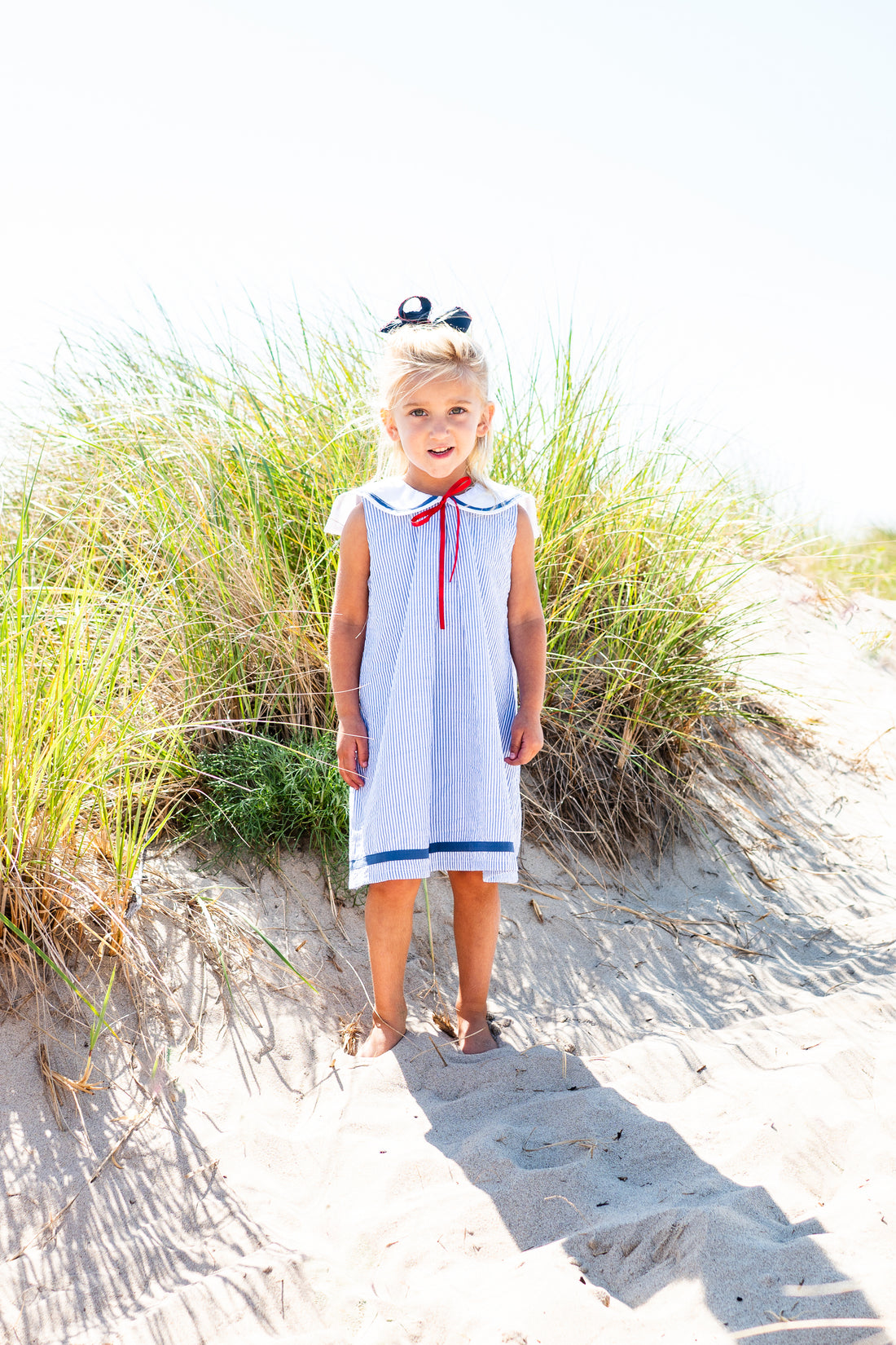 Young girl in a white dress with red and blue accents standing on sandy dunes with grass.