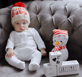 Baby in a white outfit and red hat sitting next to a snowman figurine on a gray couch.