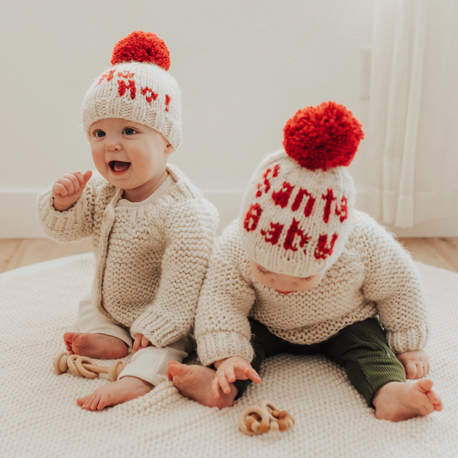 Two babies wearing knitted hats with red pom-poms and text, sitting on a light-colored blanket.