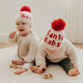 Two babies wearing knitted hats with red pom-poms and text, sitting on a light-colored blanket.