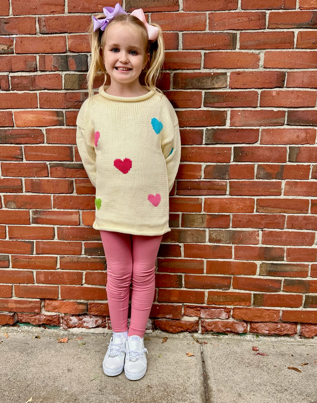 Young girl wearing a sweater with colorful heart patterns against a brick wall.