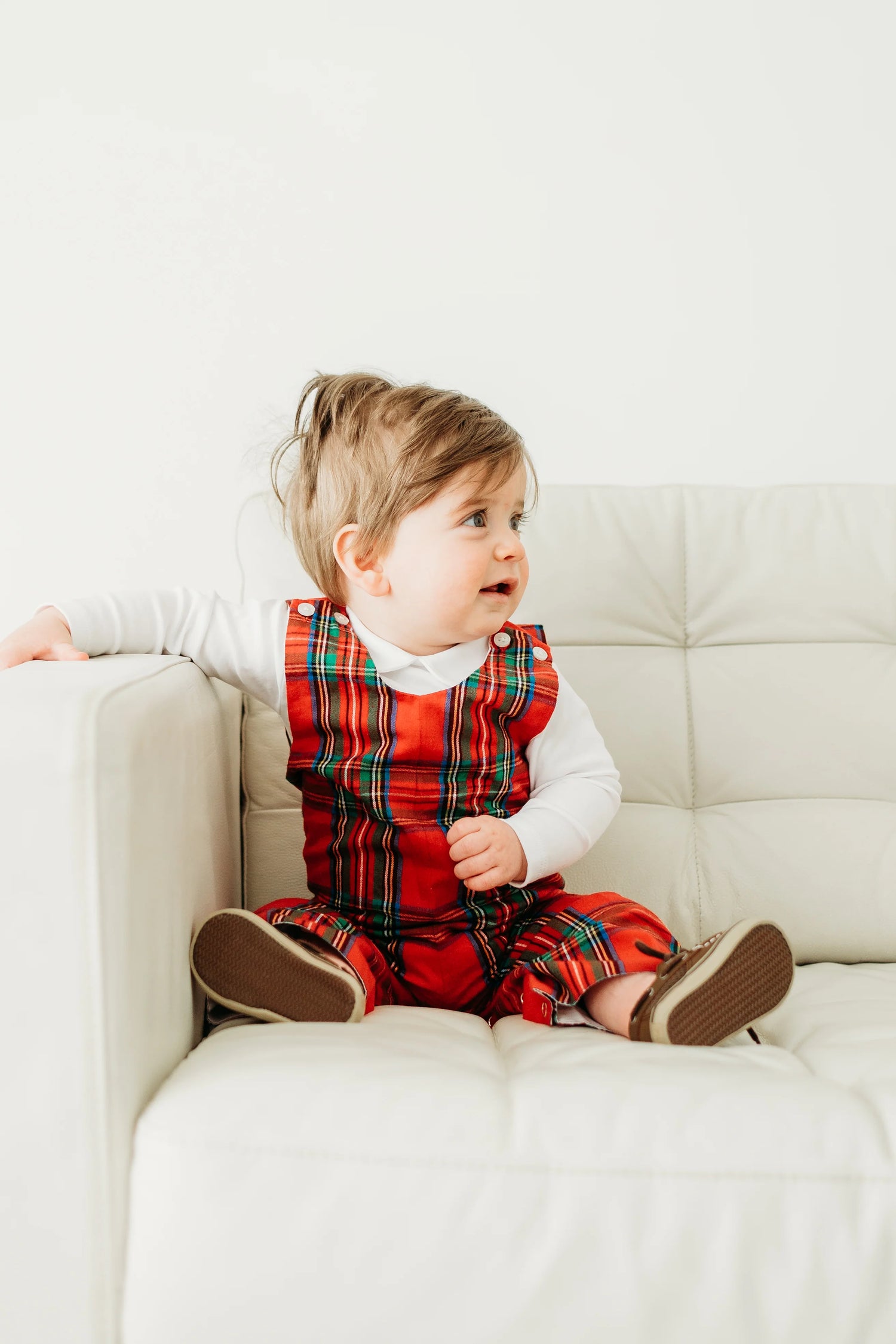 Child wearing a red plaid outfit sitting on a white couch.
