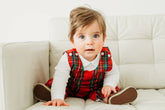 Baby wearing a red plaid outfit sitting on a white couch.