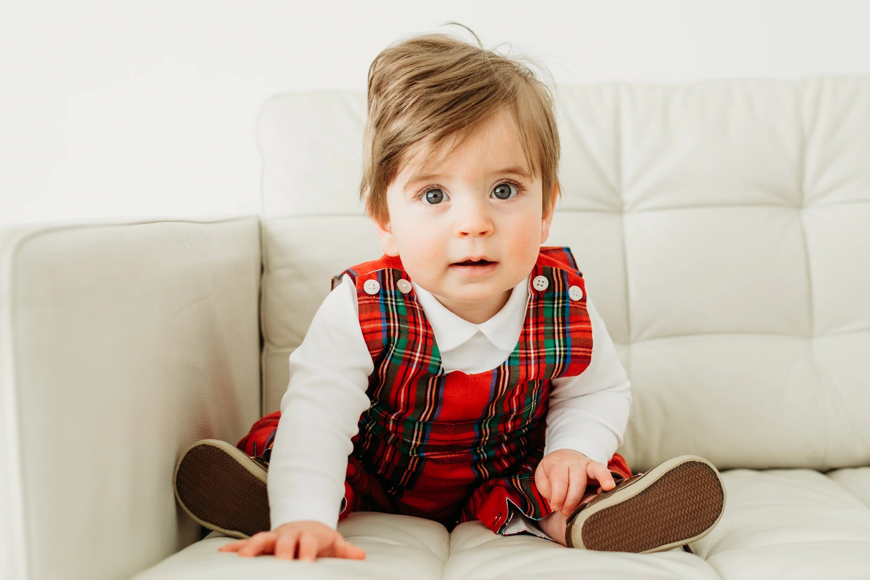 Baby wearing a red plaid outfit sitting on a white couch.