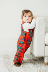 Child wearing a red plaid outfit standing on a carpeted floor.