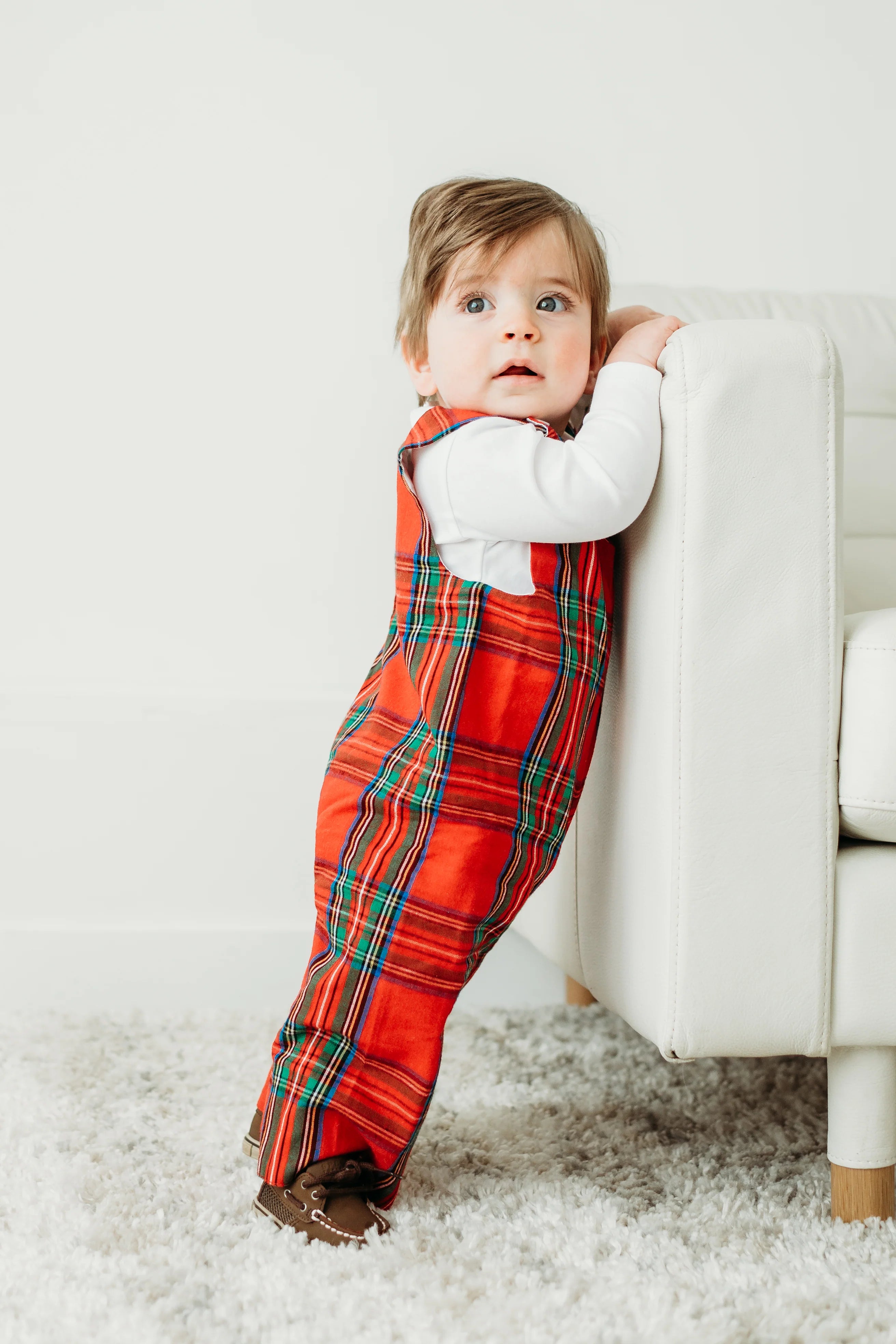 Child wearing a red plaid outfit standing on a carpeted floor.