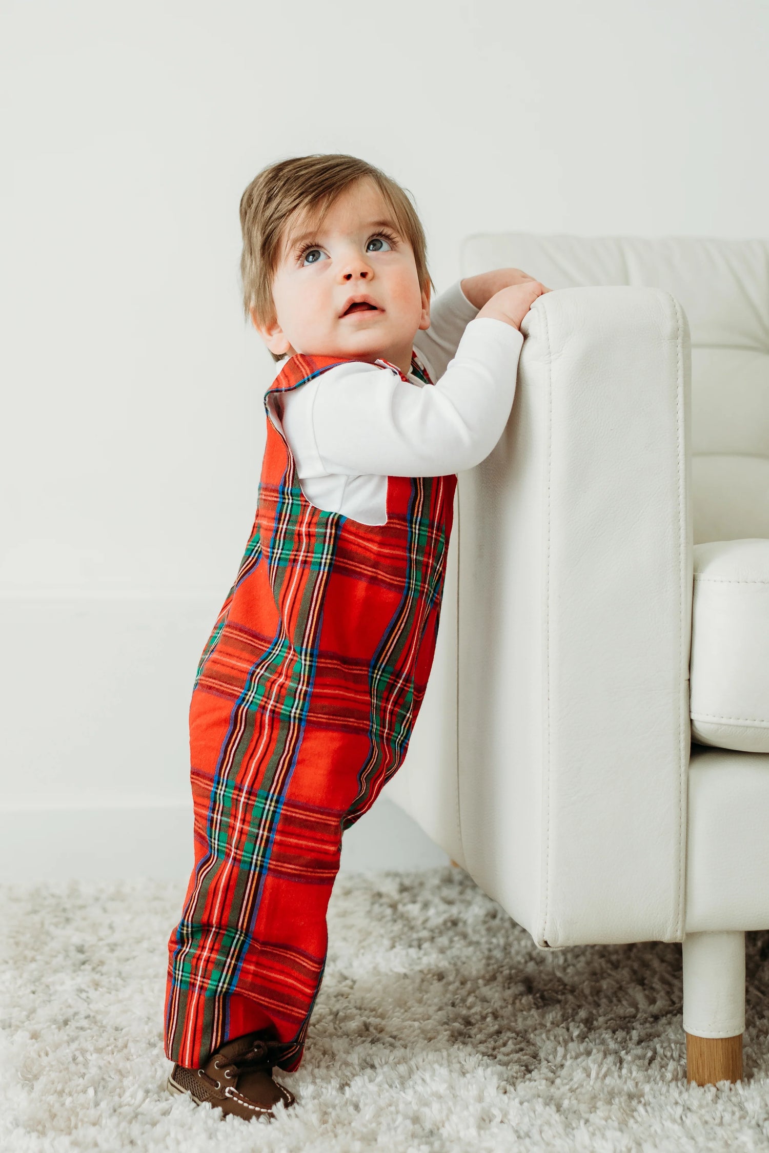 Child wearing red plaid pants and a white shirt standing on a white carpet next to a white couch.